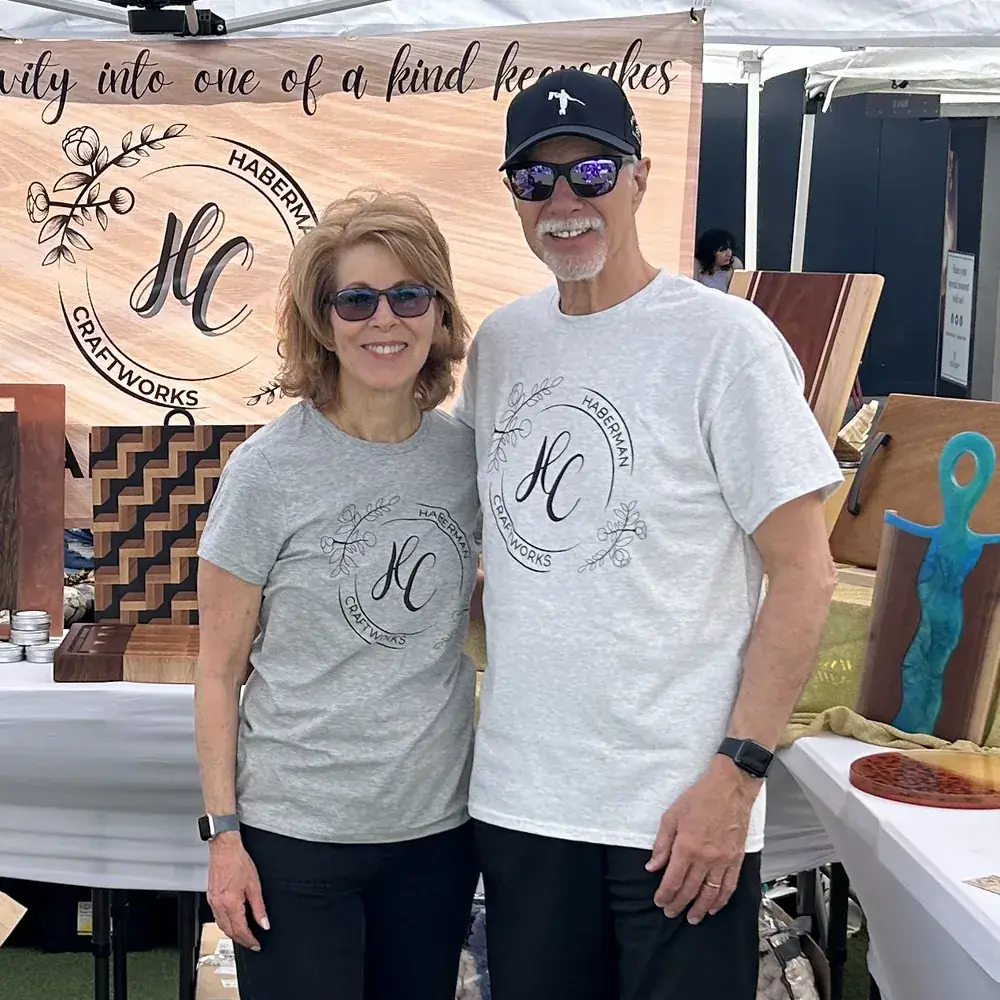 Michael and Tricia Haberman smiling side by side at their market booth, with handcrafted wood and resin boards on display behind them